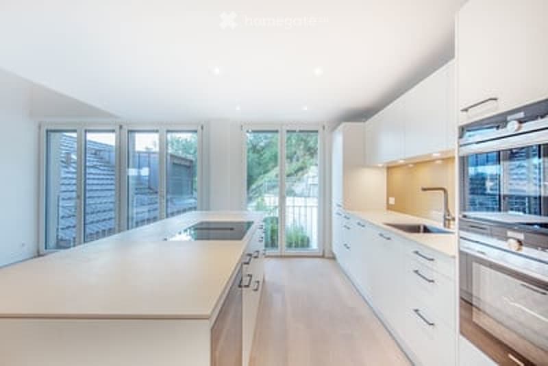 Modern kitchen with white cabinets, a central island, and stainless steel appliances. Large windows provide natural light and access to a balcony.