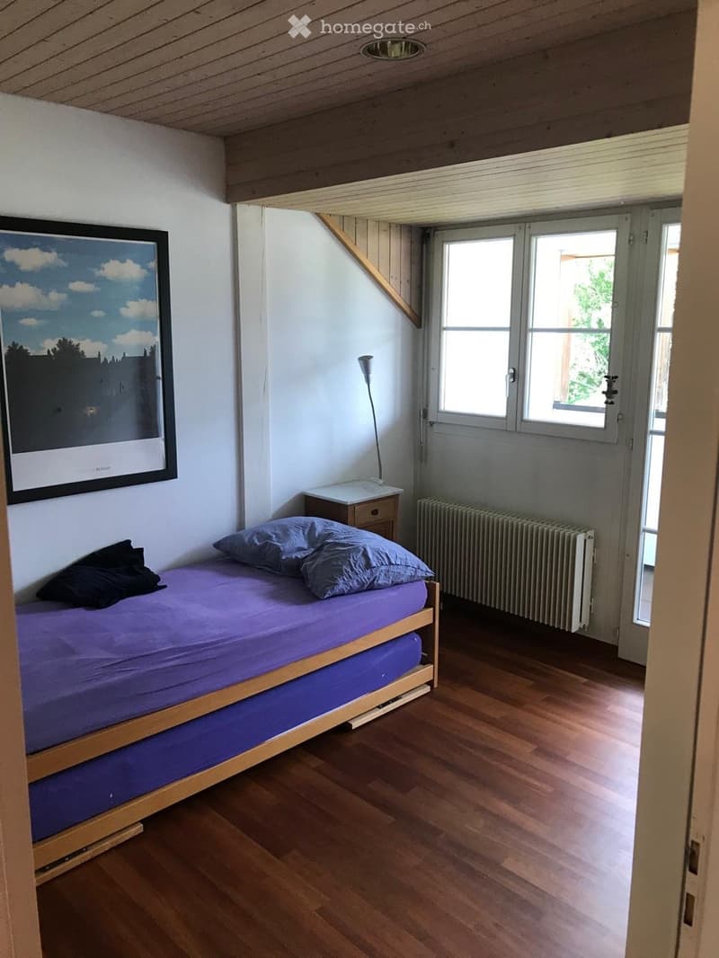 Cozy attic bedroom with wooden slanted ceilings, a single bed with purple bedding, a nightstand with a lamp, a radiator, and a large window offering natural light.