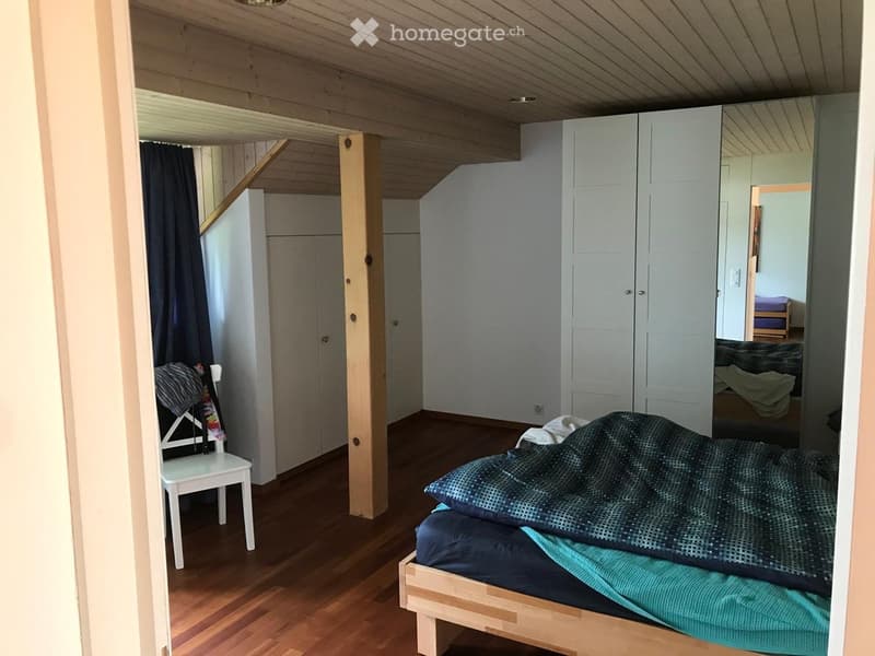 Cozy bedroom with wooden floors, a bed with a dark blue blanket, a white chair, and a large wardrobe. The room has exposed wooden beams on the ceiling and a staircase leading to another level.
