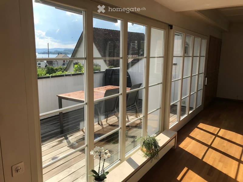 Bright balcony with large glass windows, wooden floor, table and chairs, potted plants, scenic view of water and mountains.