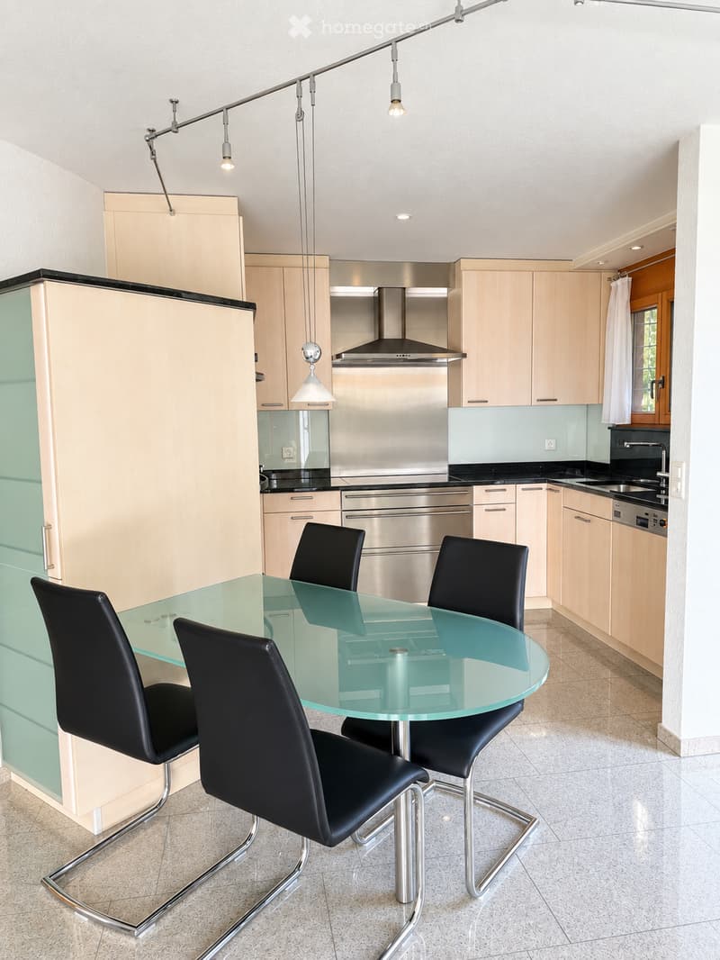 Modern kitchen with light wood cabinets, stainless steel appliances, a glass dining table with four black chairs, and a range hood.