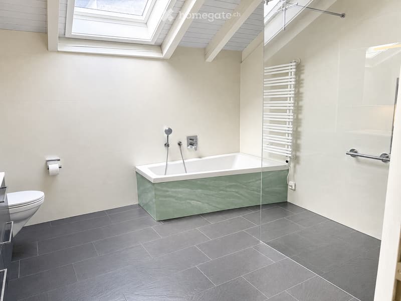 Modern bathroom with a green marble bathtub, toilet, and a radiator. The room features grey tiles on the floor and light-colored walls.
