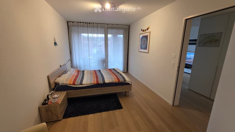 Modern bedroom with wooden flooring, a bed with striped bedding, a nightstand with a lamp, a picture frame on the wall, and a large window with curtains.