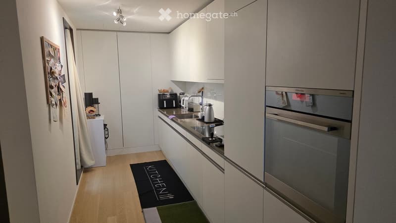 Modern kitchen with white cabinets, stainless steel appliances, wooden flooring, and a black rug with the word "KITCHEN" written on it.