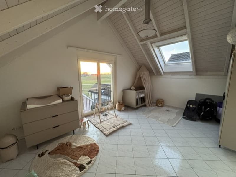 Bright, airy room with slanted wooden ceiling and skylight. Features a changing table, baby chair, crib, play area with toys, and a balcony access.