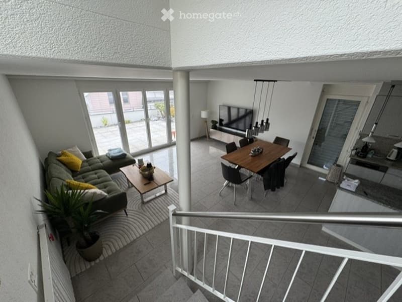 Open-plan living and dining area with modern furniture, including a grey sofa, coffee table, dining table with chairs, and a television on a stand. The space features large windows and a staircase leading to an upper level.