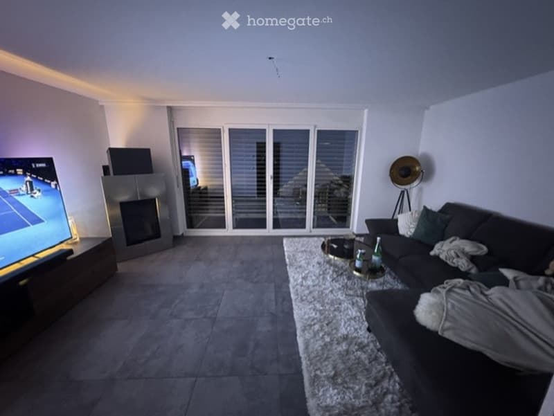Modern living room with large sliding glass doors leading to a balcony, a dark grey sofa, a coffee table with drinks, a television, and a fireplace.