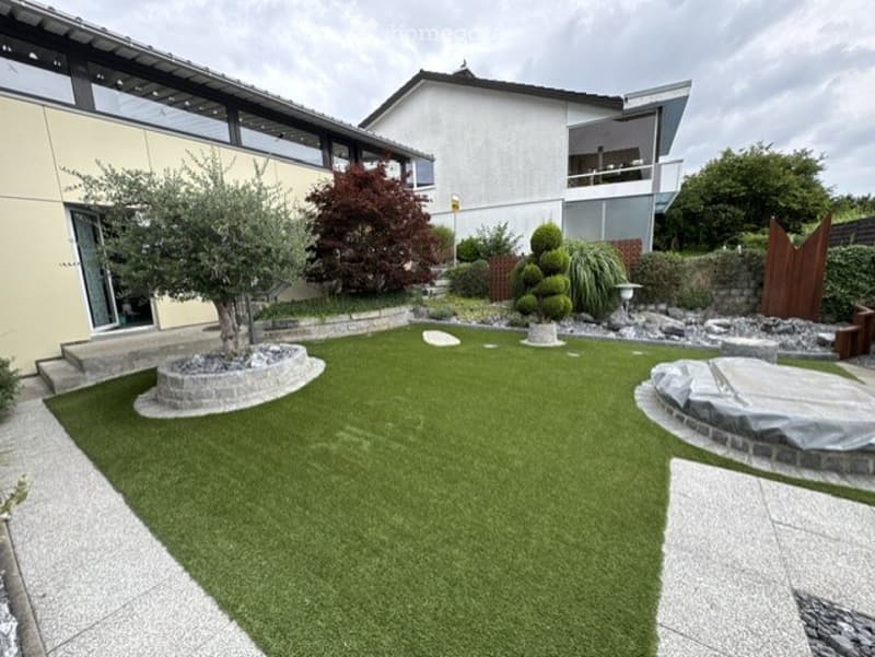 Well-maintained garden with artificial grass, small trees, shrubs, and decorative stone elements. The house in the background has a modern design with large windows and a balcony.