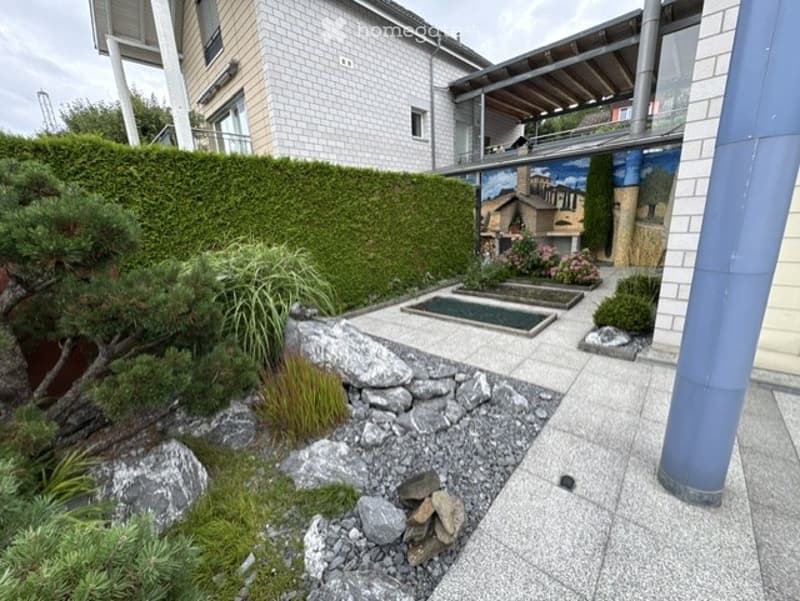 Well-maintained garden area with a small water feature, neatly trimmed hedges, rocks, and various plants, adjacent to a modern building with a balcony.