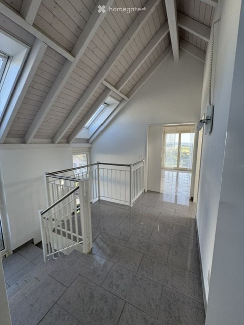 Empty room with exposed wooden beams on the ceiling, tiled floor, white walls, and a white metal staircase leading to an open area with large windows and a view.