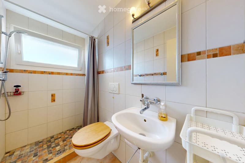 Modern bathroom with white tiles, a toilet with a wooden seat, a sink with a chrome faucet, a large mirror, a shower area with a glass door, and a small window for natural light.