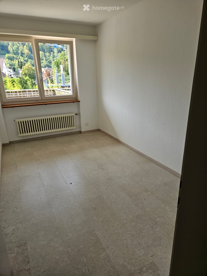 Empty room with tiled floor, white walls, radiator, and large window with view of greenery.