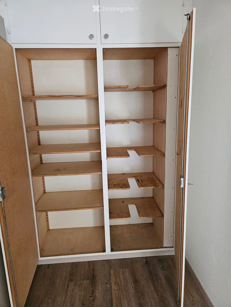 Empty wooden closet with multiple shelves and white upper cabinets, wooden floor, white walls.