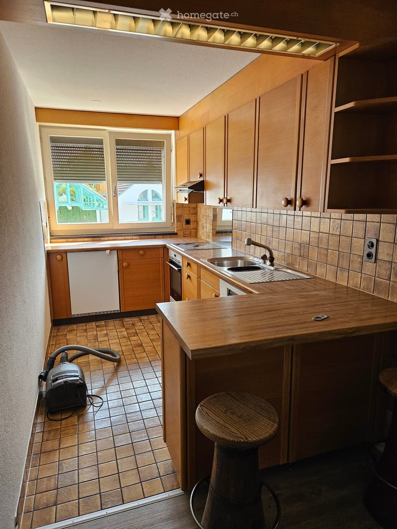 Modern kitchen with wooden cabinets, a central island, a sink, and a stool. The space features tiled flooring and a window with blinds.