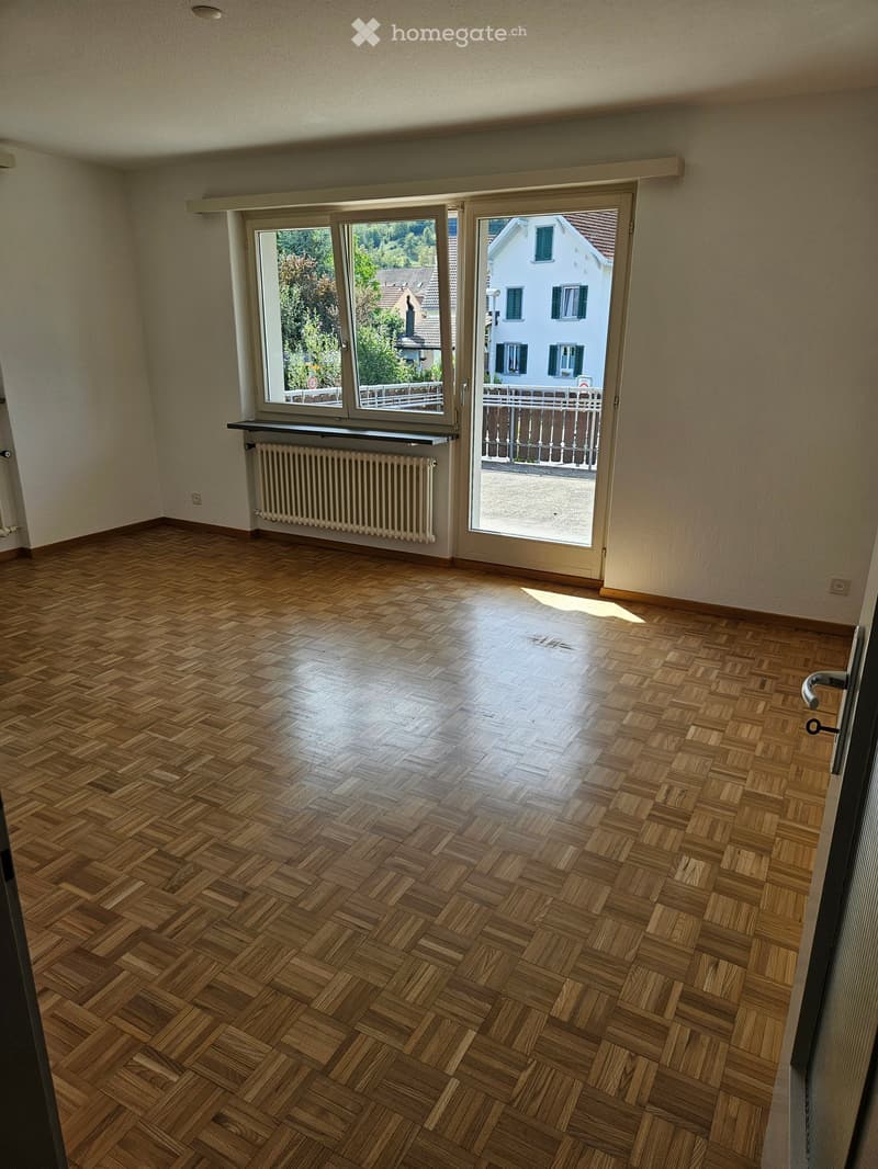 Empty room with wooden herringbone flooring, large windows, radiator, and sliding glass door leading to a balcony.