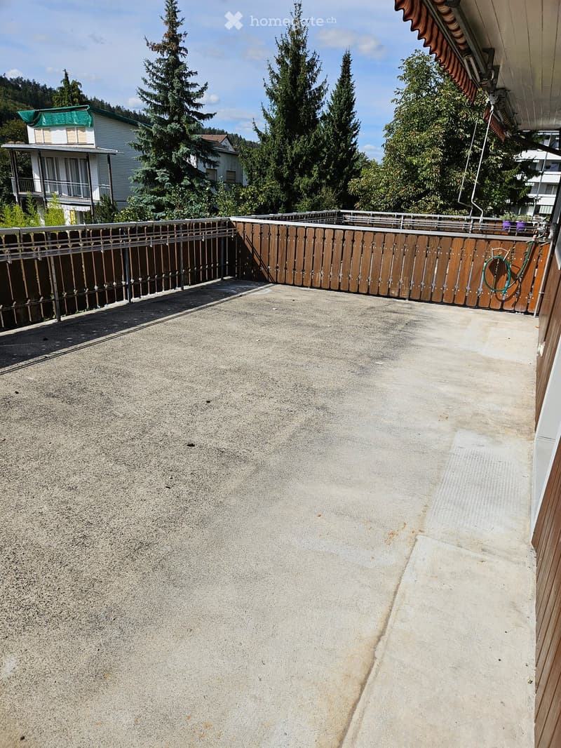 Empty concrete balcony with wooden and metal railings, surrounded by trees and neighboring buildings.