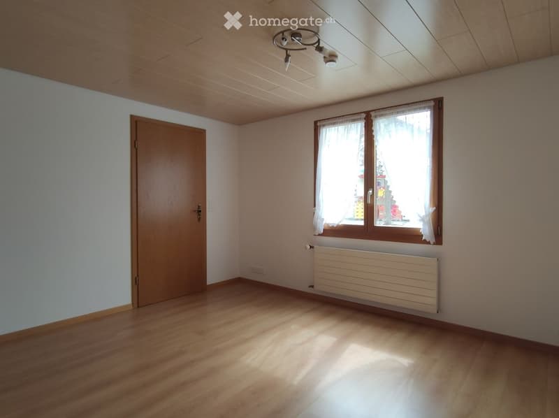 Empty room with wooden flooring, white walls, a window with white curtains, a wooden door, and a radiator below the window.