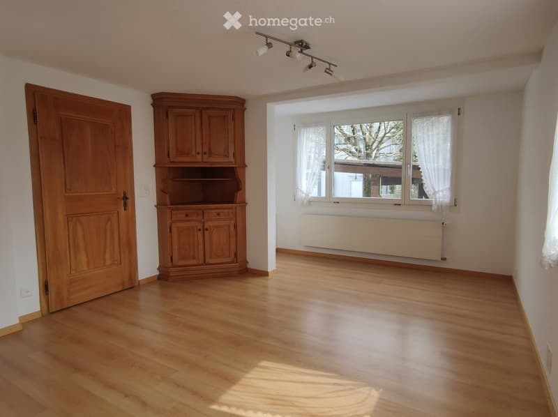 Empty room with wooden floor, large window with white curtains, wooden cabinet, and track lighting.
