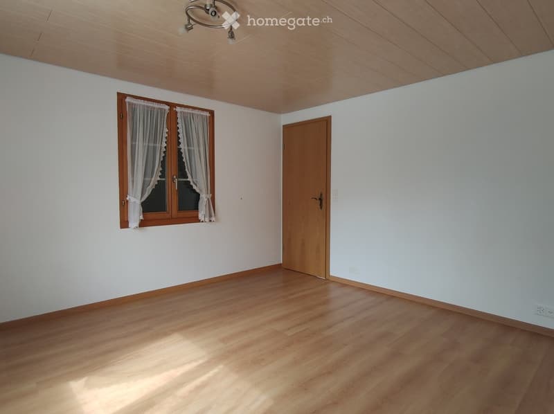 Empty room with wooden flooring, white walls, and a wooden door. A window with white curtains is visible on one wall.