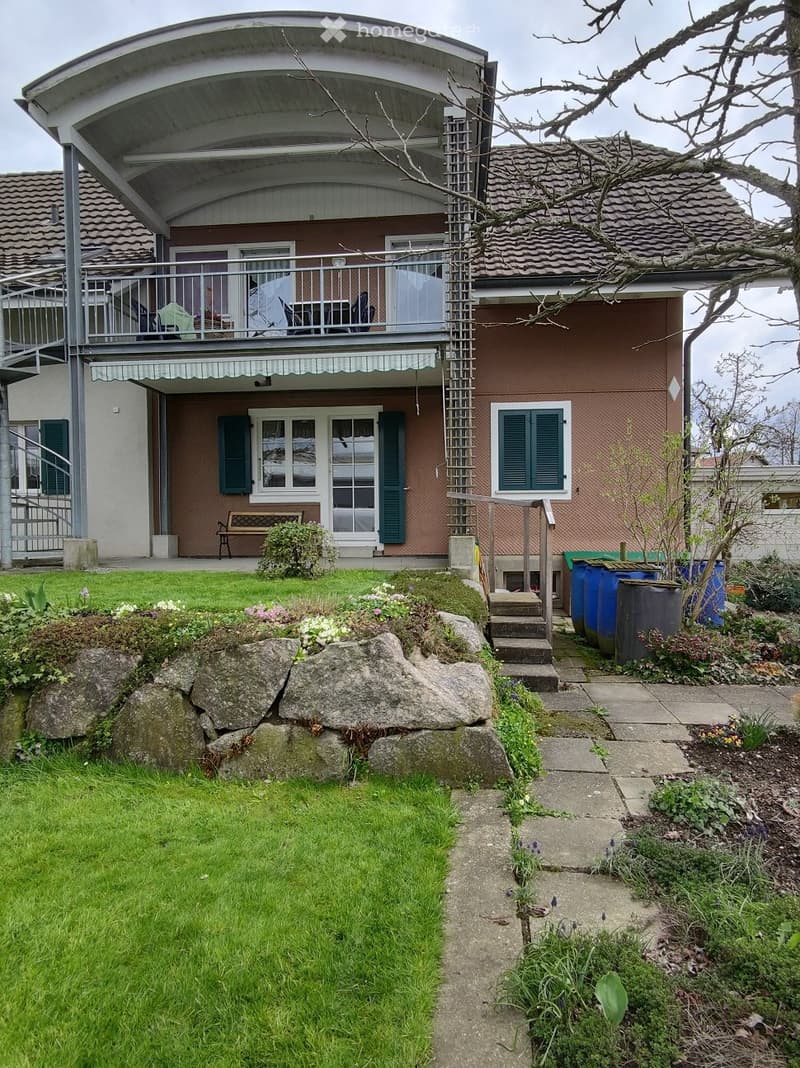 Garden area with a well-maintained lawn, stone pathway, and small garden beds. A modern house with a balcony and covered patio is visible in the background.