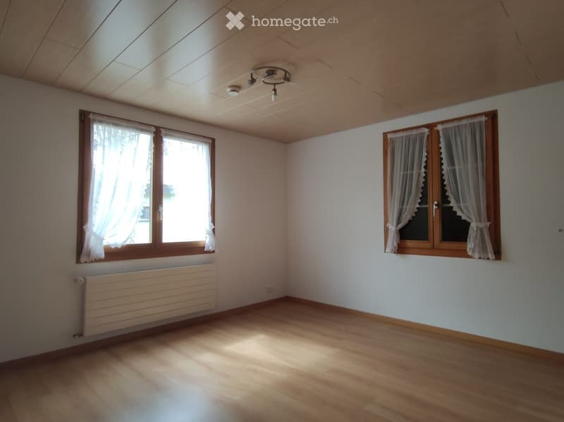 Empty room with wooden flooring, white walls, large windows with white curtains, and a radiator under one window.
