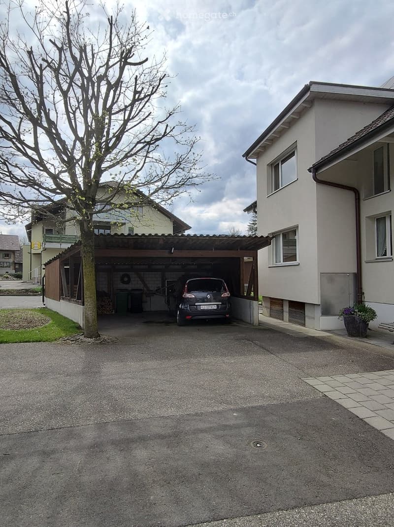 A two-story residential building with a covered garage, a parked car, and a tree in the front yard.