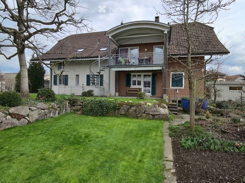Two-story house with a curved balcony, green shutters, and a well-maintained garden with a lawn and flower beds.