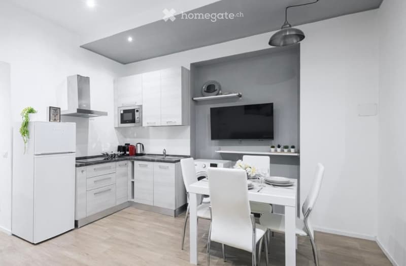 Modern, open-plan kitchen and dining area with white cabinets, stainless steel appliances, a dining table with chairs, and a television mounted on the wall.