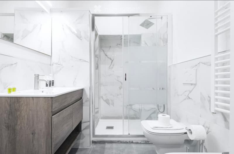 Modern bathroom with marble walls, a glass-enclosed shower, a wooden vanity with a sink, and a toilet with a towel and toilet paper.
