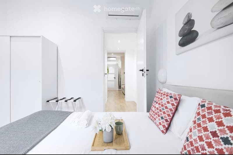 Modern, minimalist bedroom with a grey duvet, decorative pillows, a wooden tray with a vase of white flowers and a cup, a wardrobe with a mirror, and a hallway leading to another room.
