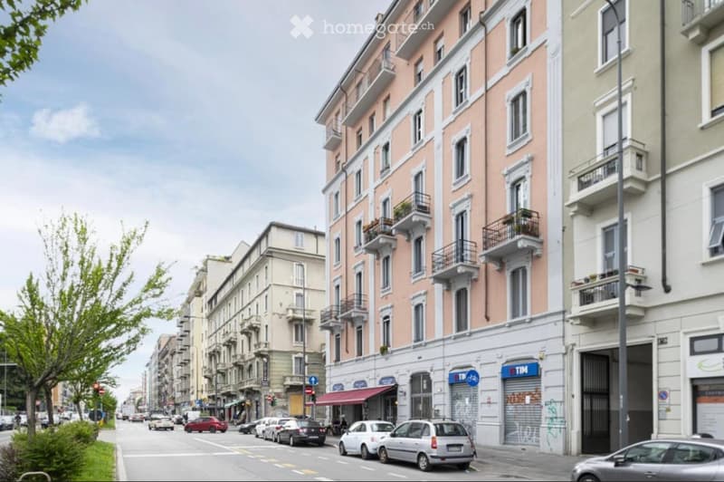 A street view of a multi-story building with balconies and awnings, surrounded by parked cars and trees.