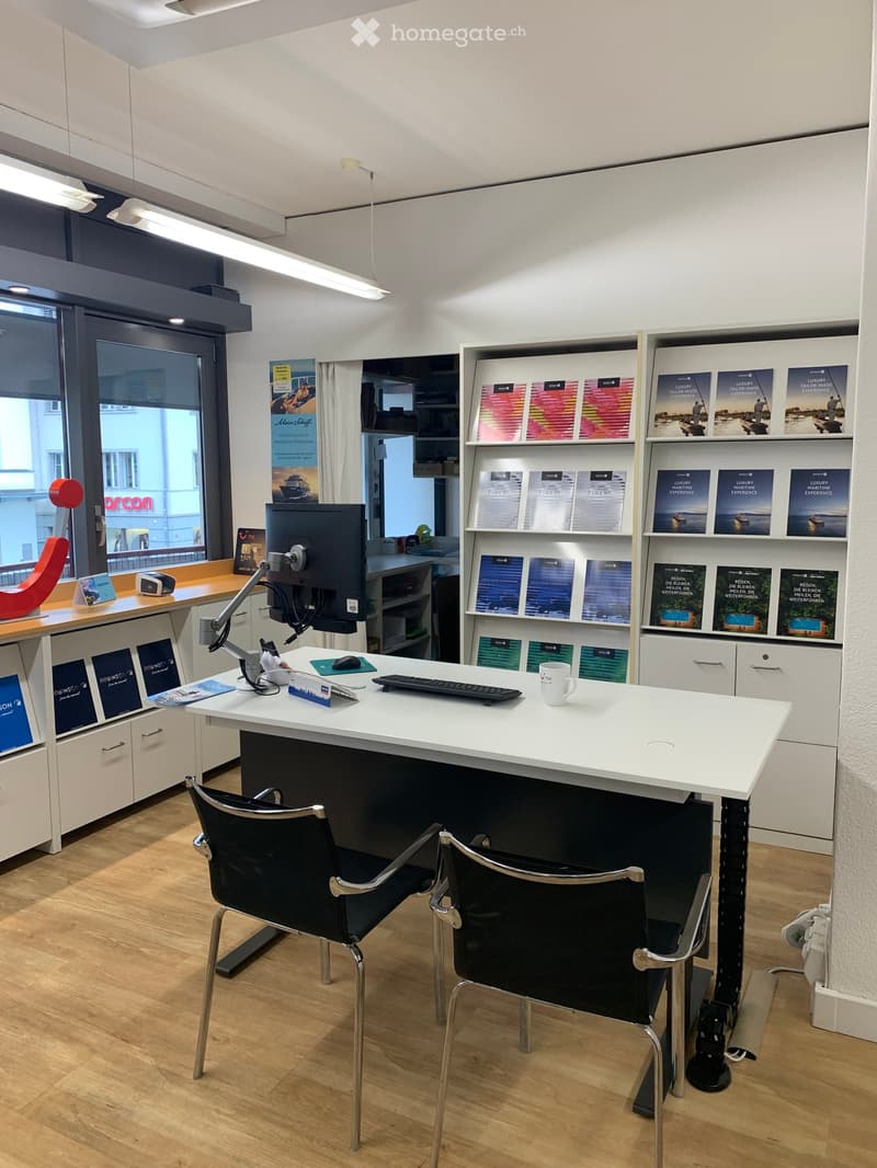 Modern office space with a white desk, two black chairs, computer setup, and shelves holding brochures and promotional materials.