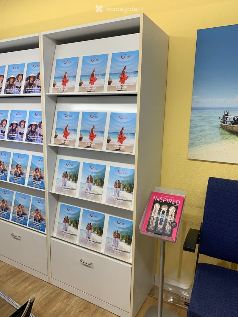A display area with brochures on shelves and a chair, featuring travel brochures and a poster of a beach scene.