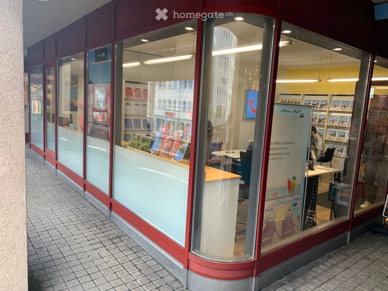 Modern storefront with large glass windows, red and white facade, interior visible with desks, chairs, and display boards.