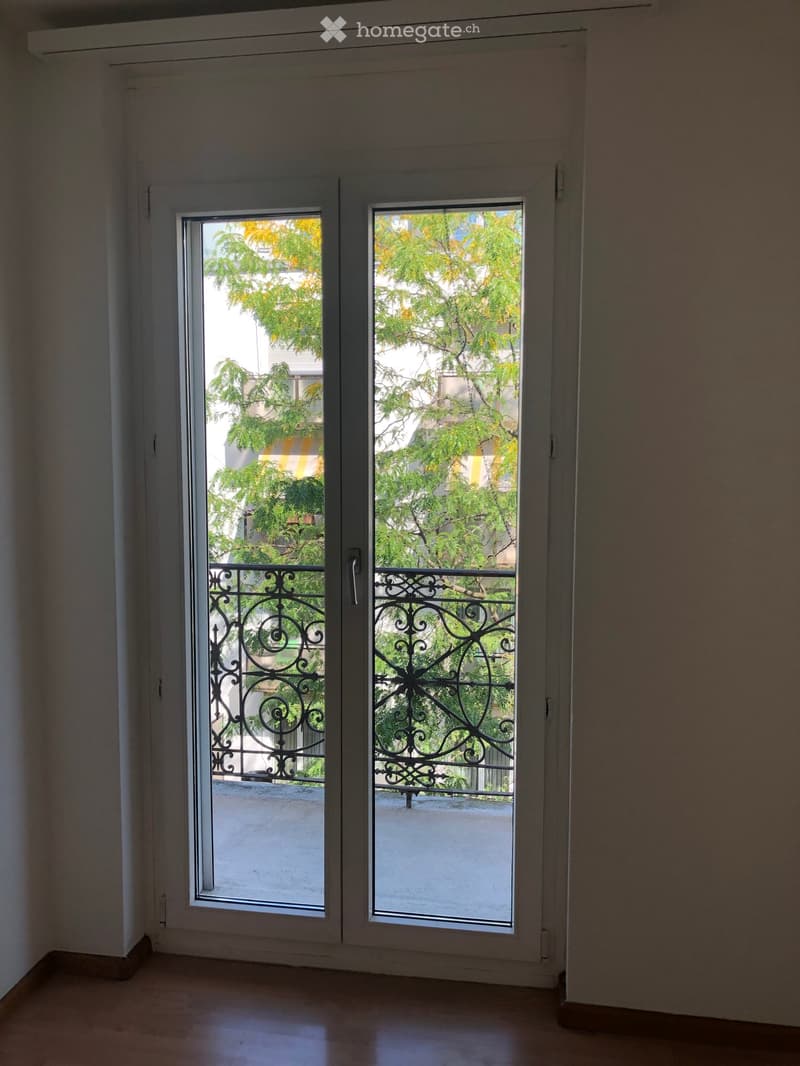 Empty room with large sliding glass doors leading to a balcony with metal railings and greenery visible outside.