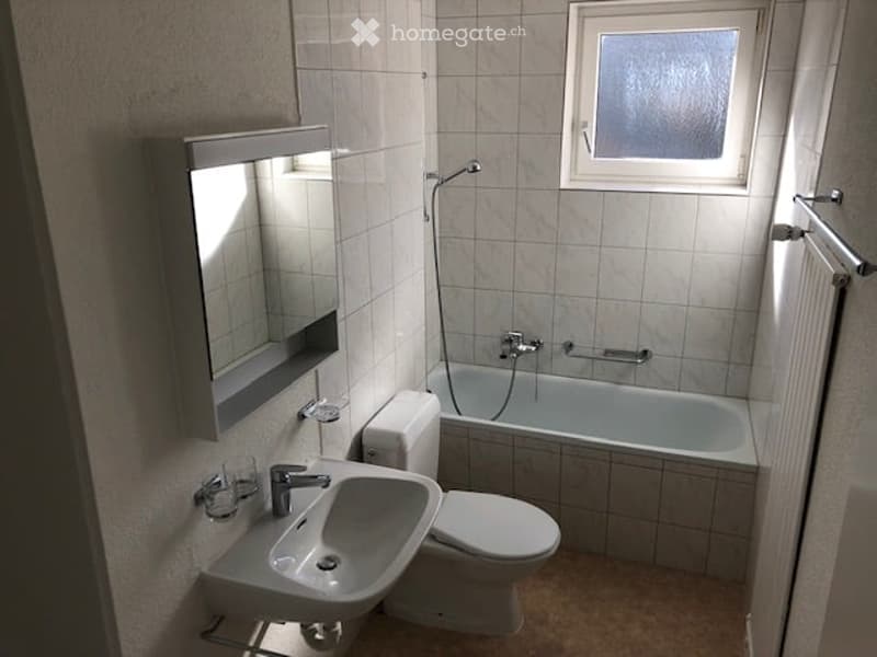 Modern bathroom with white tiles, a sink, toilet, bathtub, and a window above the tub.