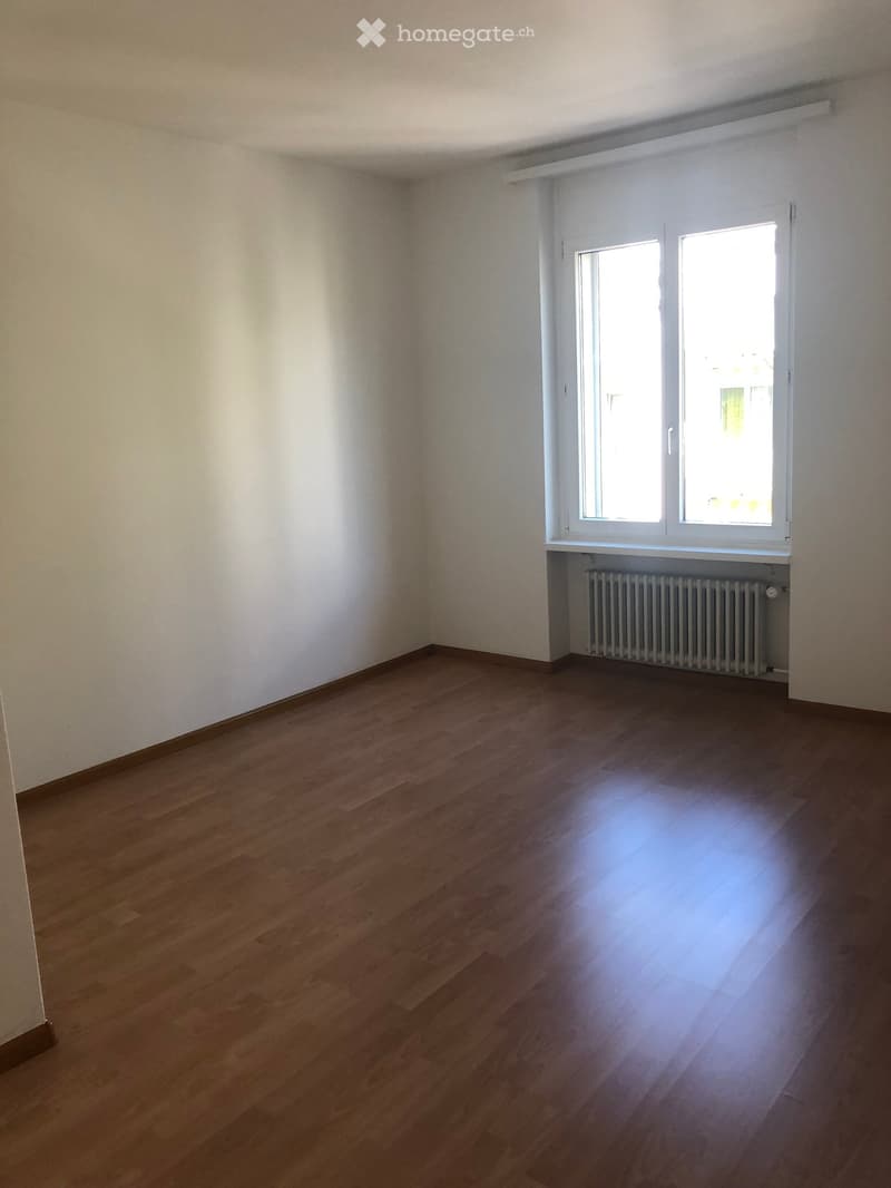 Empty room with wooden flooring, white walls, and a window with white frame. A radiator is visible below the window.