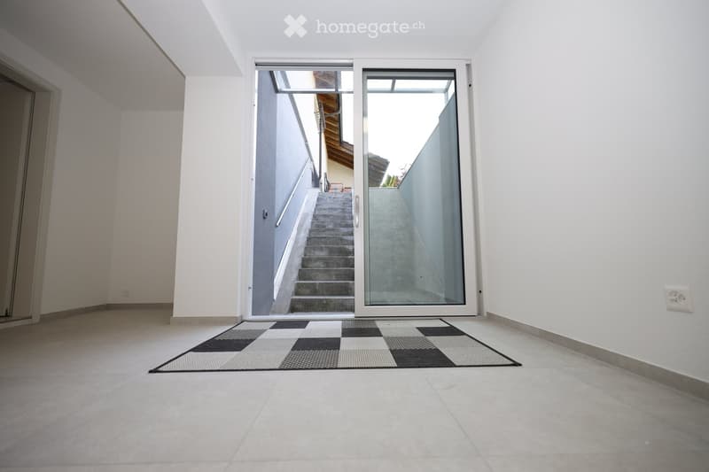 Empty hallway with white walls, tiled floor, and a checkered doormat leading to a staircase with a glass door.