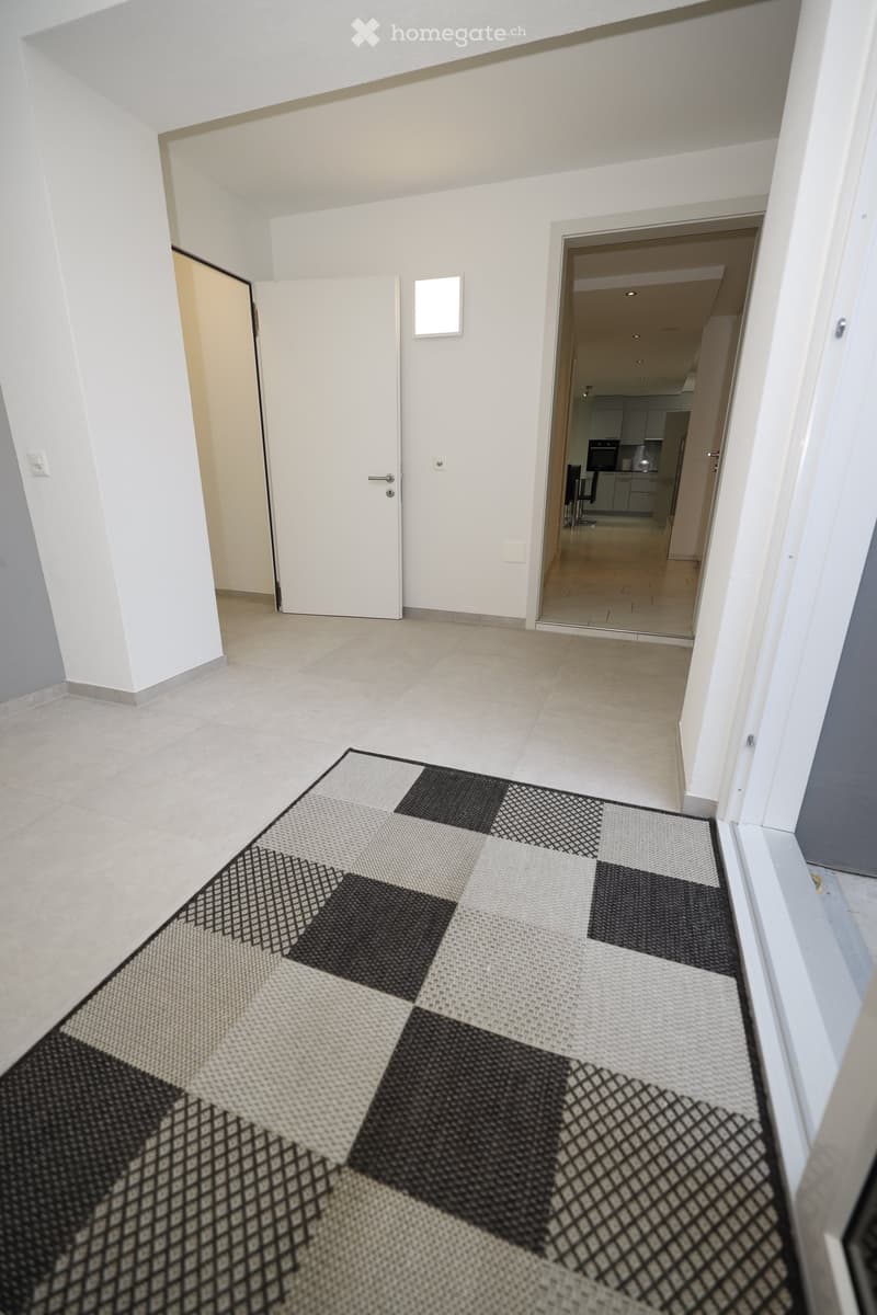 Empty hallway with tiled floor, white walls, and doors leading to other rooms. A black and white checkered rug is placed on the floor.