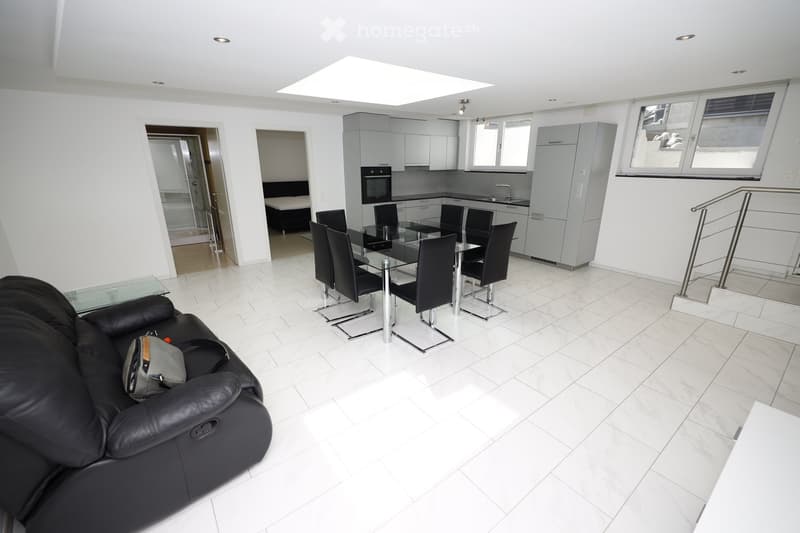 Modern, well-lit living and dining area with white tiled flooring, a black leather sofa, a glass dining table with six black chairs, and a fitted kitchen with grey cabinets, an oven, and a sink.