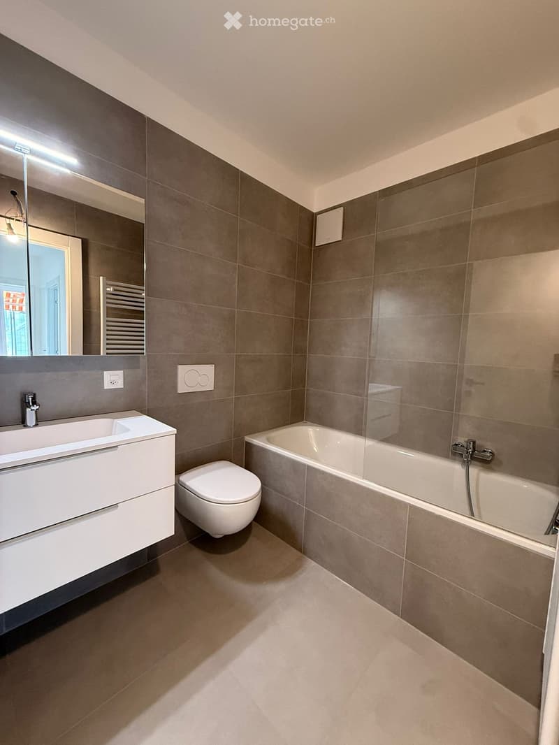 Modern bathroom with grey tiled walls, a white sink, toilet, bathtub, and a large mirror above the sink.