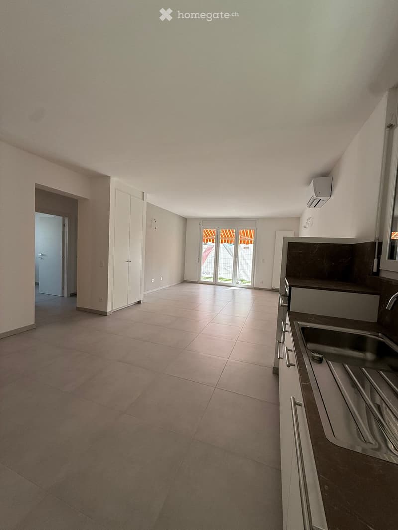 Empty room with tiled floor, white walls, and a modern kitchen area featuring a sink and cabinets. A sliding glass door leads to an outdoor area.