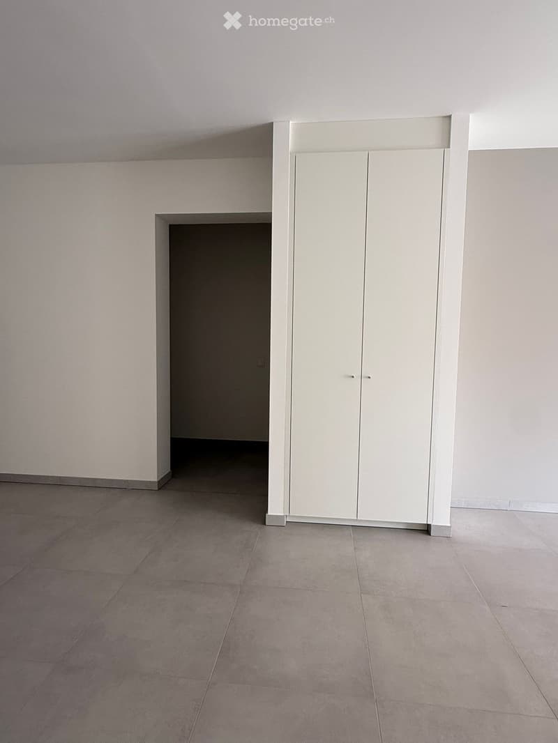 Empty room with light grey tiled floor and white walls, featuring a built-in white cabinet with double doors and a small open shelf section.