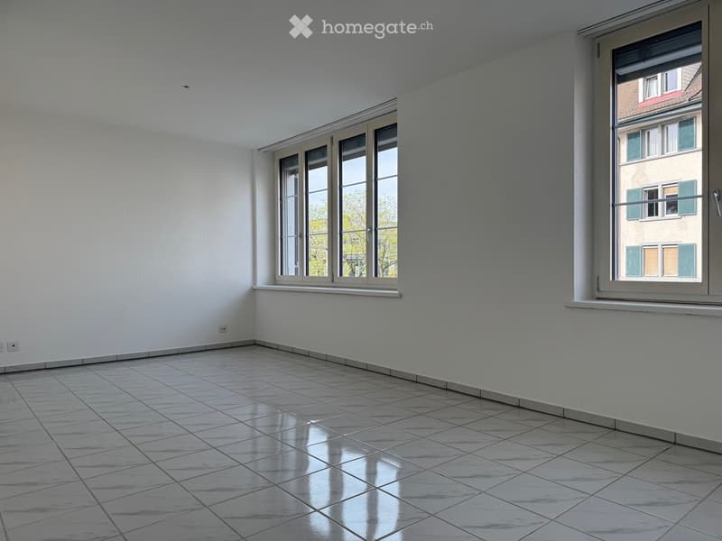 Empty room with white walls, tiled floor, and large windows offering a view of neighboring buildings.