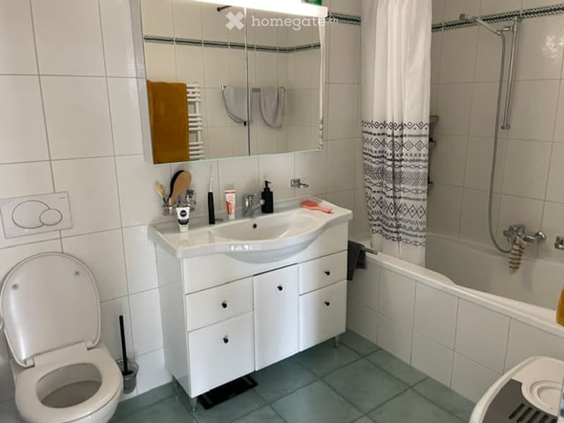 Modern bathroom with white tiles, a white sink cabinet, a large mirror, a toilet, and a bathtub with a shower curtain.