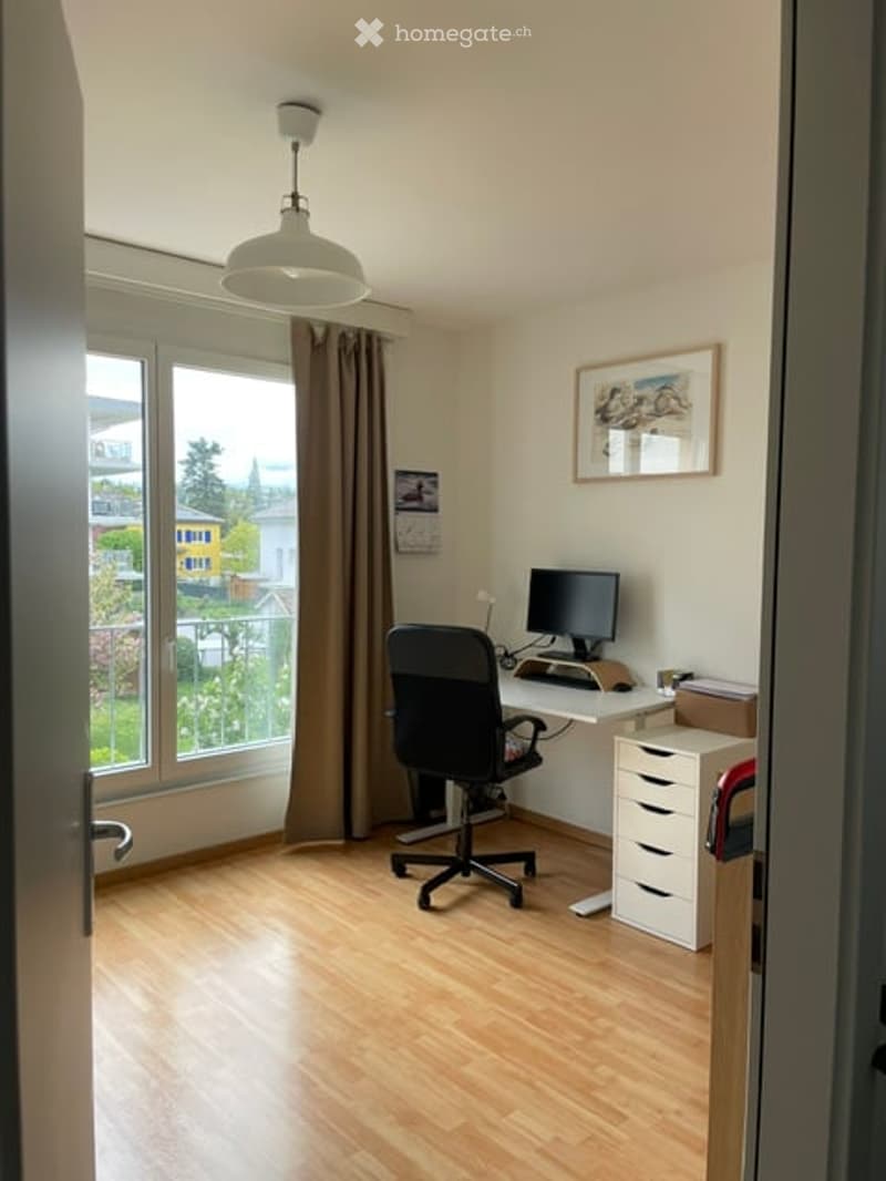 Bright, modern home office with wooden flooring, large windows offering a view, a desk with a computer, a black office chair, a white storage unit, and a hanging lamp.