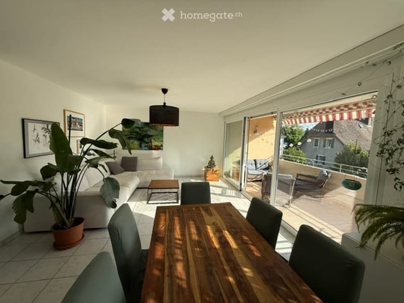 Bright living and dining area with large windows and sliding doors leading to a balcony. Features a white sofa, coffee table, dining table with chairs, floor lamp, and decorative plant.