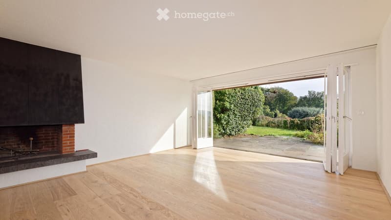 Empty room with wooden flooring, white walls, and a brick fireplace. Large sliding glass doors open to a garden.