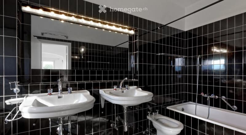 Modern bathroom with black tiled walls, two white sinks, a bathtub, and a toilet. Features include a large mirror with integrated lighting and a sleek faucet design.