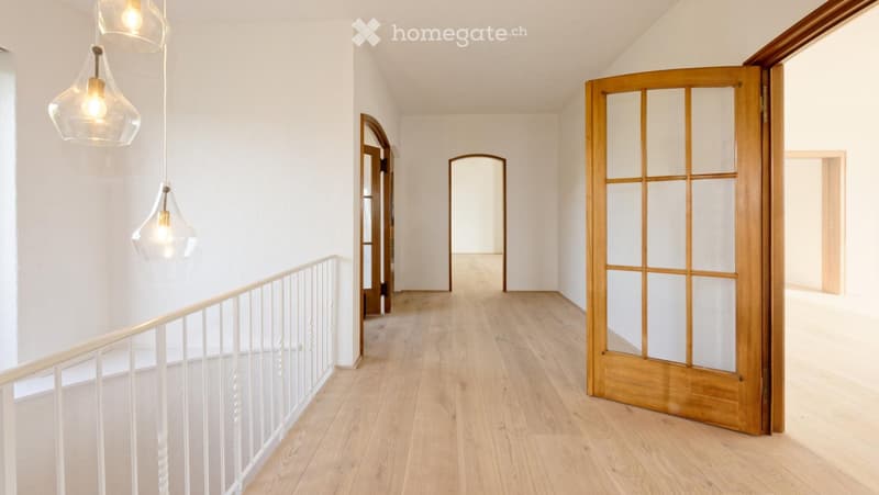 Bright, modern hallway with light wooden flooring, white walls, and pendant lights. Features include a white railing on the left and several doorways leading to other rooms.